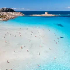 Turkusowa woda, biały piasek i kamienna wieża na wysepce – to jedne z najpiękniejszych plaż w Europie. Ludzie kąpią się i spacerują po płytkiej wodzie.