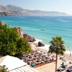 Costa del Sol w lutym: słoneczna plaża z palmami, turkusowe morze i góry w tle. Ludzie odpoczywają pod parasolami.