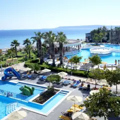 Słoneczna plaża i baseny w Ialyssos na Rodos. Dzieci zjeżdżają ze zjeżdżalni, dorośli odpoczywają na leżakach.