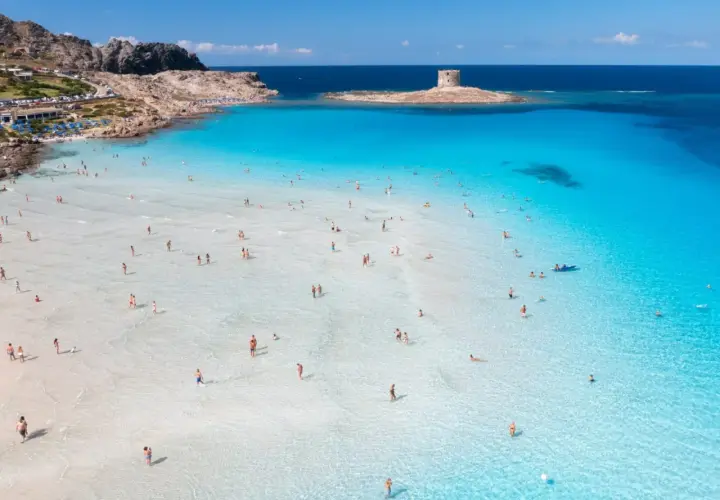 Turkusowa woda, biały piasek i kamienna wieża na wysepce – to jedne z najpiękniejszych plaż w Europie. Ludzie kąpią się i spacerują po płytkiej wodzie.