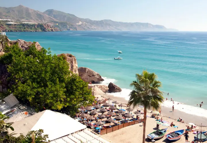Costa del Sol w lutym: słoneczna plaża z palmami, turkusowe morze i góry w tle. Ludzie odpoczywają pod parasolami.