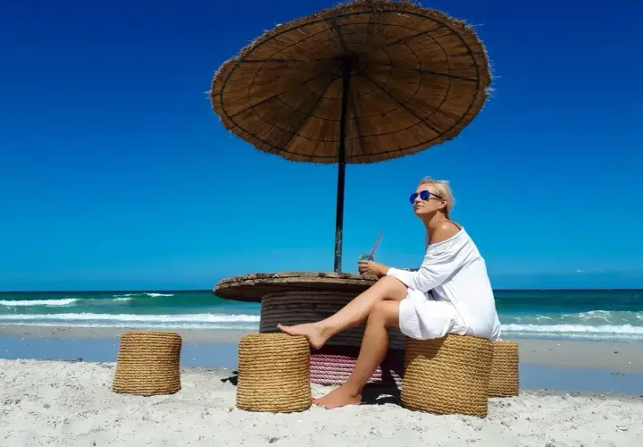 Kobieta w białej sukience siedzi pod parasolem na plaży Djerby. Uważaj na słońce i upał, pijąc orzeźwiający napój.