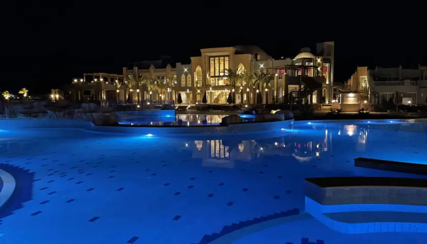 Nocny basen z niebieskim podświetleniem i luksusowym budynkiem hotelu Lazuli Marsa Alam w tle.