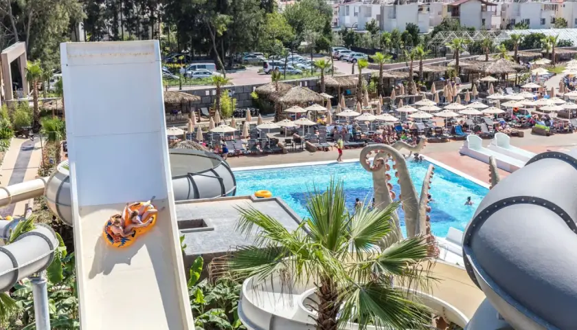 Dwie osoby zjeżdżają na pontonie ze zjeżdżalni wodnej w Atlantique Holiday Club. W tle basen i leżaki.