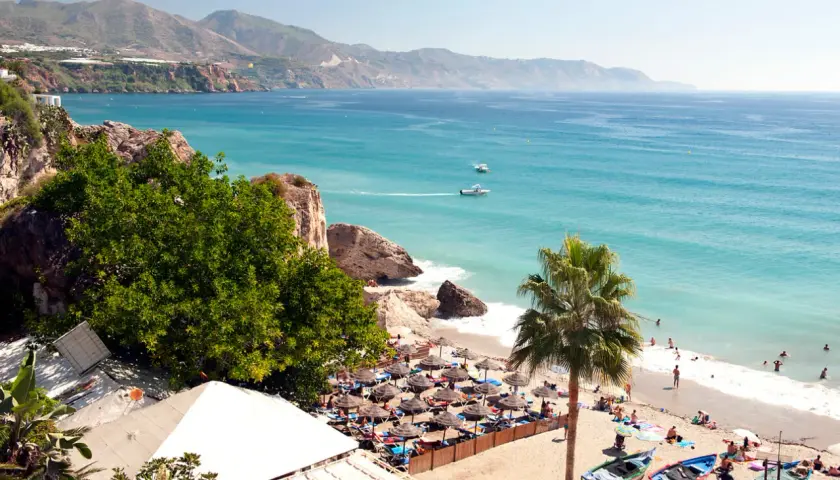 Costa del Sol w lutym: słoneczna plaża z palmami, turkusowe morze i góry w tle. Ludzie odpoczywają pod parasolami.