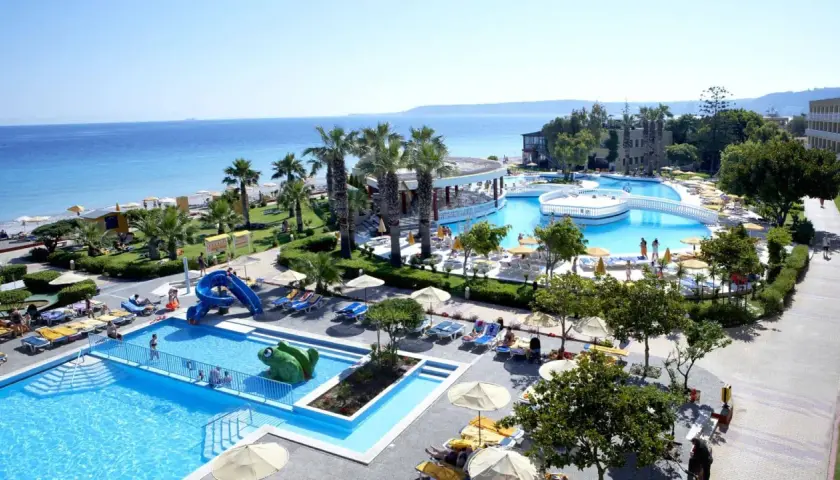 Słoneczna plaża i baseny w Ialyssos na Rodos. Dzieci zjeżdżają ze zjeżdżalni, dorośli odpoczywają na leżakach.