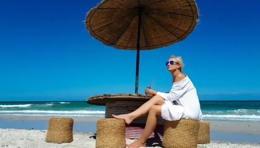 Kobieta w białej sukience siedzi pod parasolem na plaży Djerby. Uważaj na słońce i upał, pijąc orzeźwiający napój.