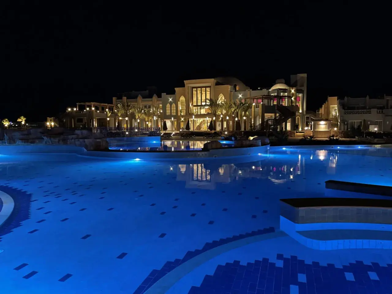 Nocny basen z niebieskim podświetleniem i luksusowym budynkiem hotelu Lazuli Marsa Alam w tle.
