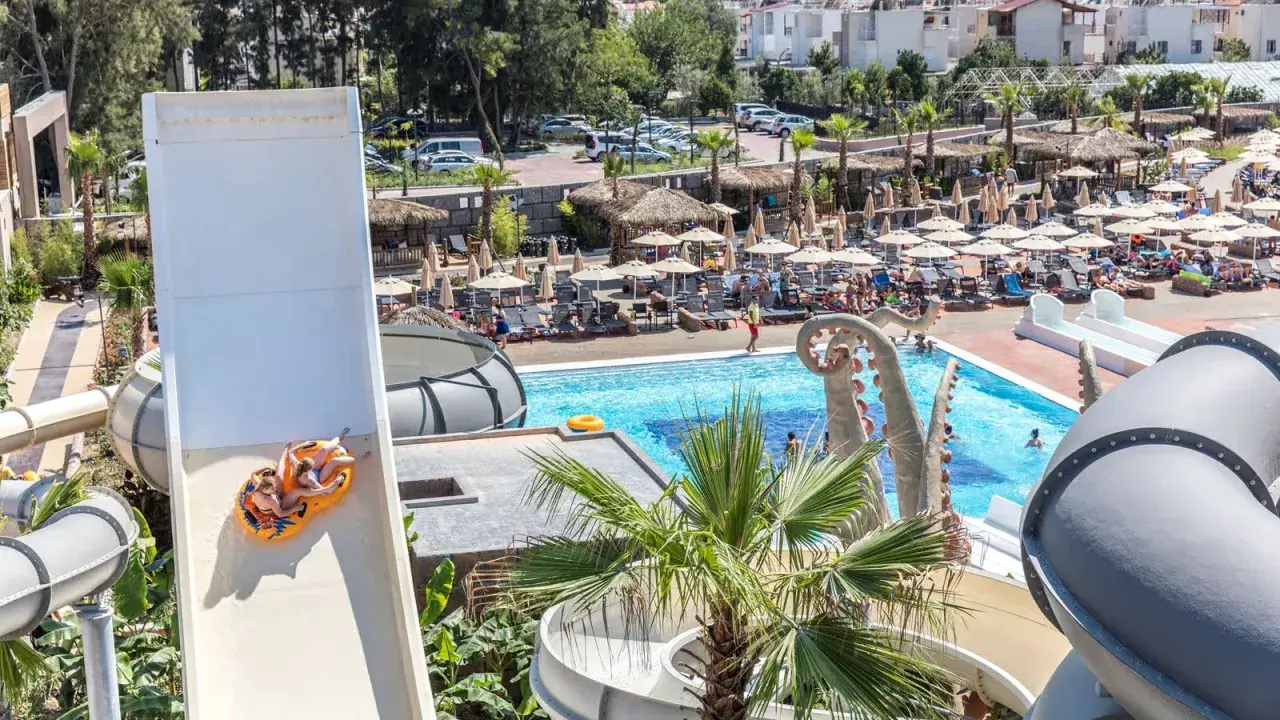 Dwie osoby zjeżdżają na pontonie ze zjeżdżalni wodnej w Atlantique Holiday Club. W tle basen i leżaki.