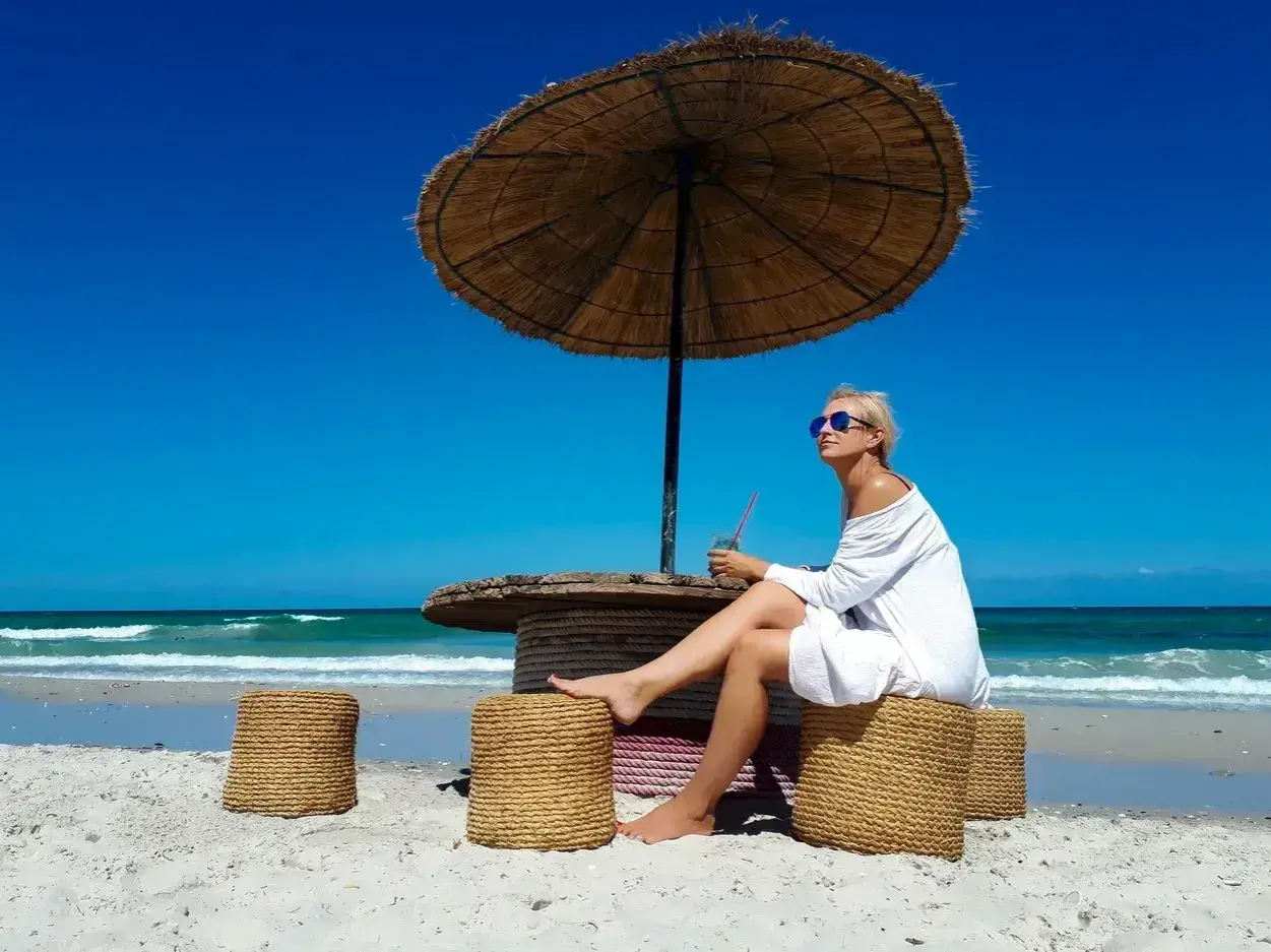 Kobieta w białej sukience siedzi pod parasolem na plaży Djerby. Uważaj na słońce i upał, pijąc orzeźwiający napój.