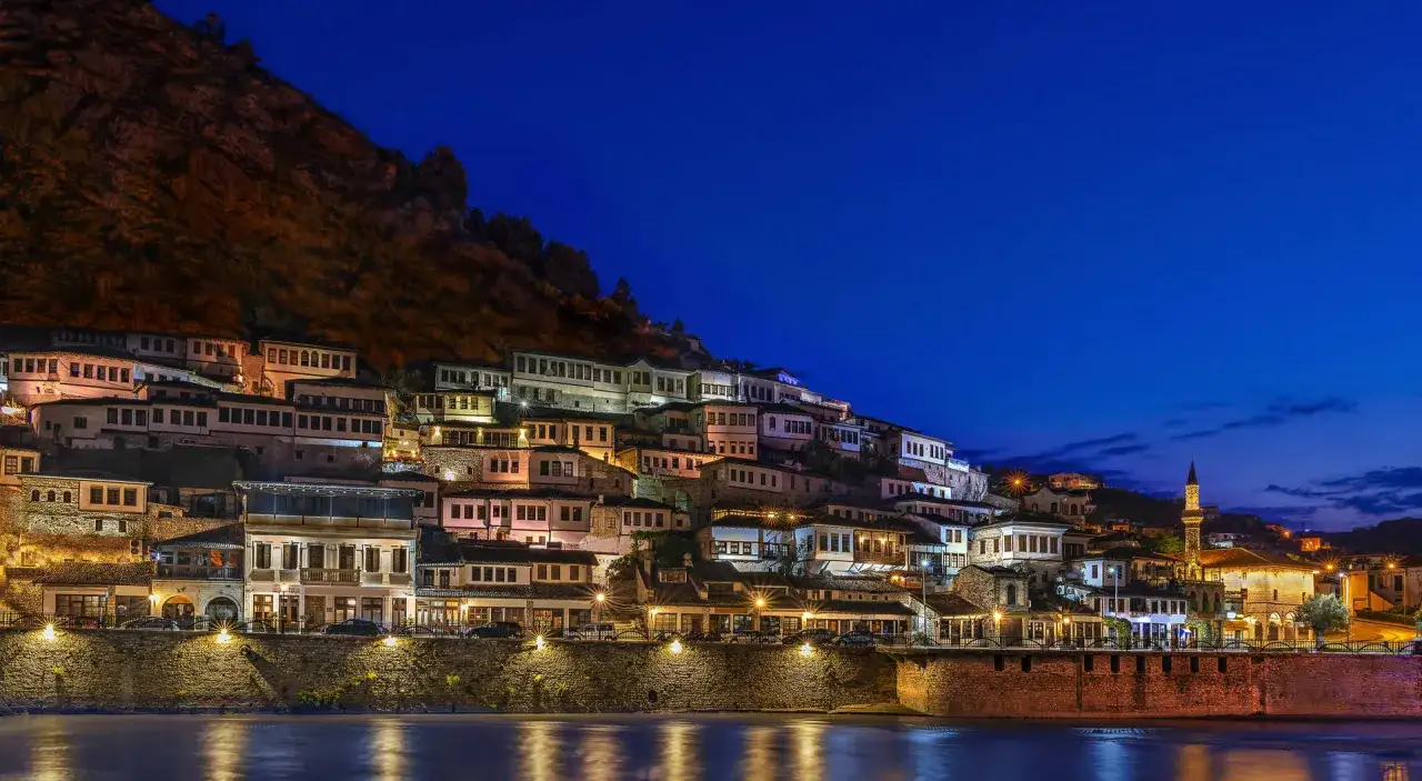 Albania, styczeń. Nocą oświetlone domy na zboczu góry w Berat, odbijające się w rzece.