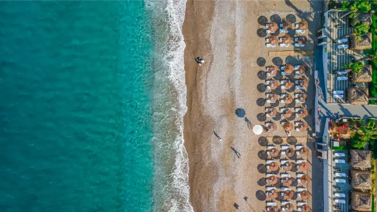 Luksusowa plaża w Okurcalar, Turcja. Turkusowe morze, piaszczysty brzeg z leżakami i parasolami, kilka osób spaceruje.