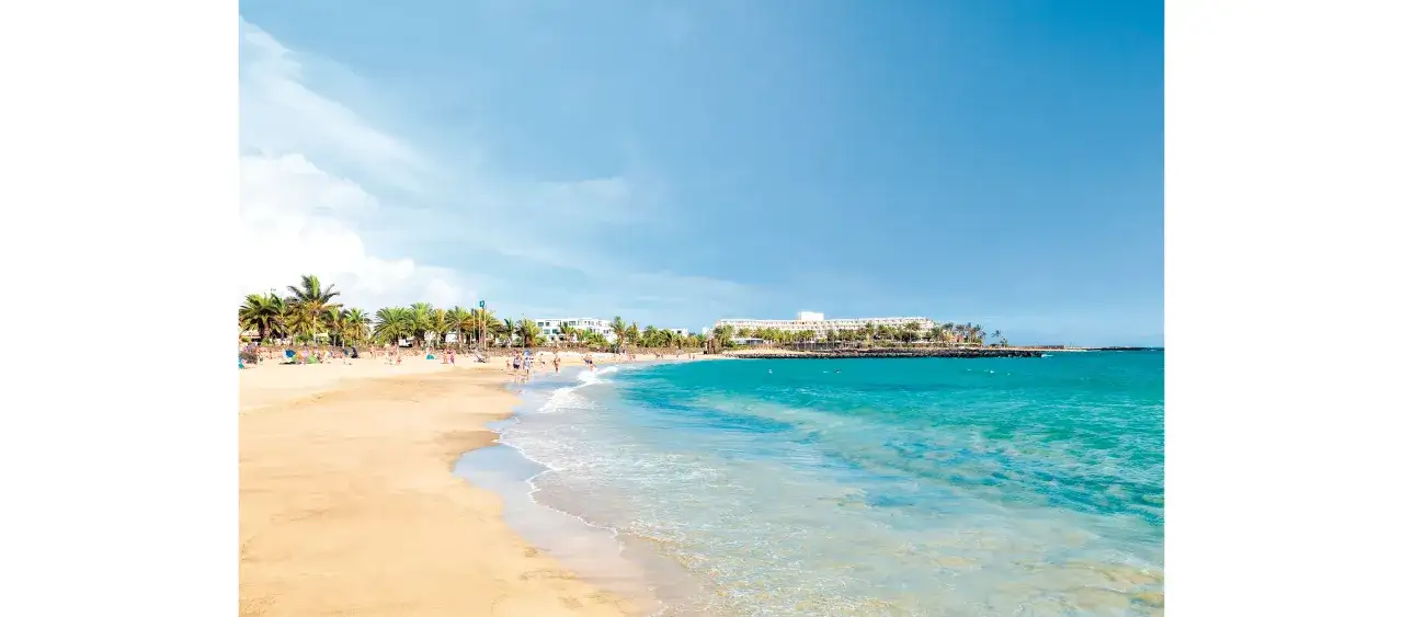 Plaża z palmami i turkusową wodą, w tle hotel Barceló Lanzarote Active Resort.