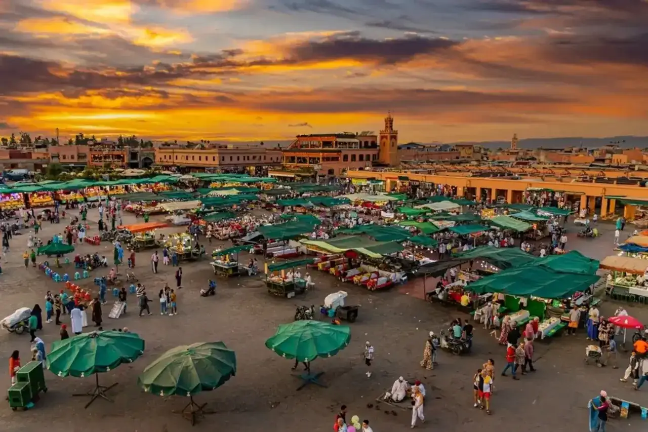 Tętniący życiem plac w Marrakeszu o zachodzie słońca. Tłumy ludzi spacerują między straganami pod zielonymi parasolami. Czy w Maroko jest bezpiecznie? Tak, to miejsce pełne życia i kultury.
