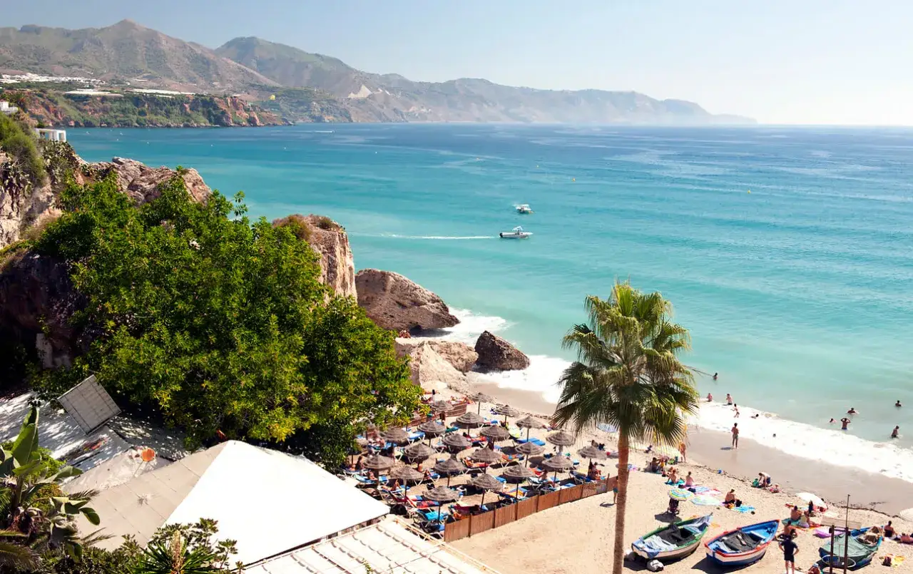 Costa del Sol w lutym: słoneczna plaża z palmami, turkusowe morze i góry w tle. Ludzie odpoczywają pod parasolami.