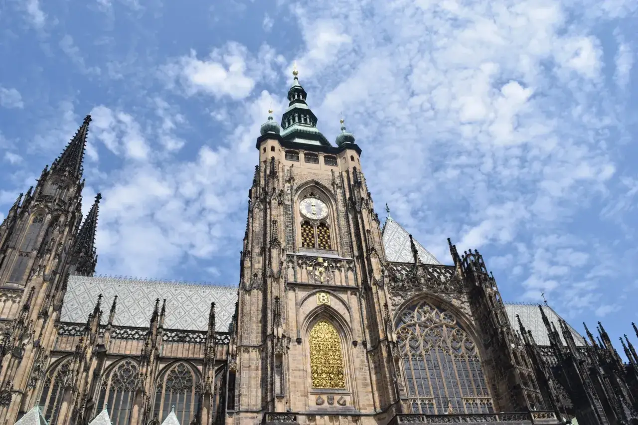 Majestatyczny zamek w Pradze, z dominującą wieżą zegarową i strzelistymi wieżyczkami, wznosi się na tle błękitnego nieba z chmurami.