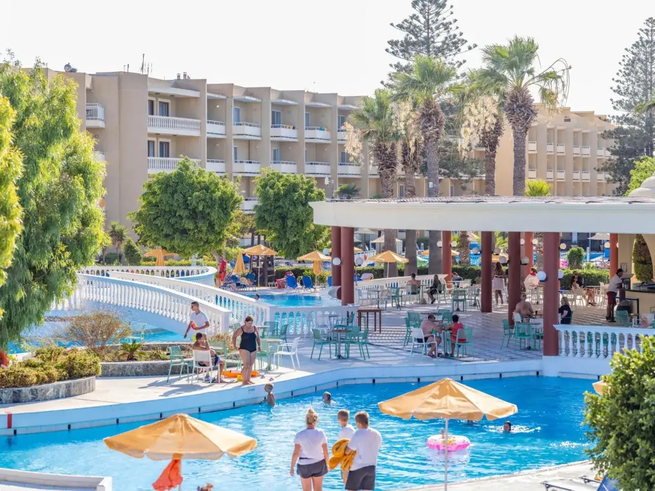 Słoneczny dzień w hotelu w Ialyssos na Rodos. Ludzie relaksują się przy basenie, pod palmami.