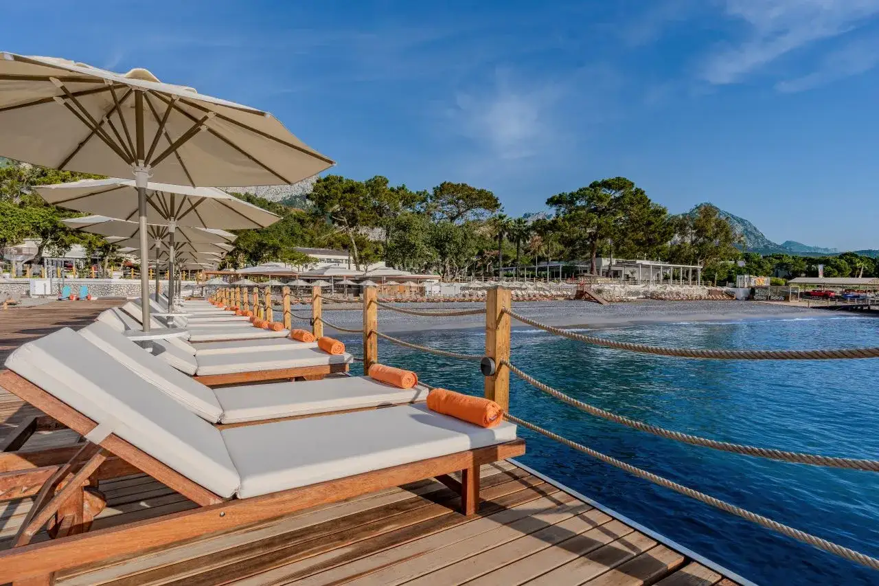 Rząd leżaków z pomarańczowymi ręcznikami i parasolami na drewnianym molo, z widokiem na plażę i morze w Bergiz Hotels & Resorts Kemer.