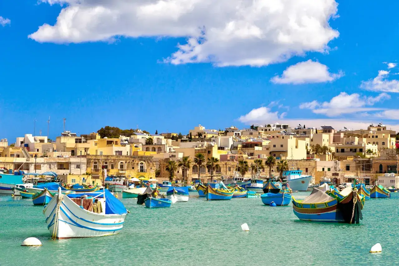 Malta w czerwcu: błękitne niebo, białe chmury i kolorowe łodzie rybackie w zatoce. Idealna pogoda na zwiedzanie.